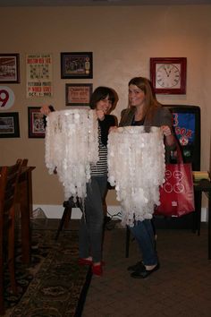 two women standing in a room holding up large white pieces of clothing that are hanging from the ceiling