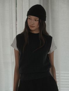 a woman standing in front of a white curtain wearing a black hat and sweater vest