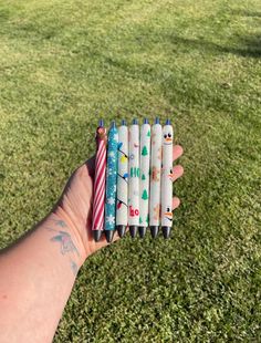 a person holding six pens in their left hand on the grass with trees and snowmen painted on them