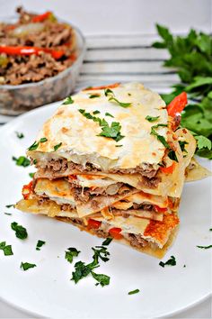 a white plate topped with layered lasagna next to a bowl of meat and veggies