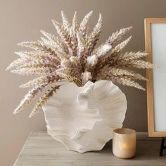 a white vase with dried plants in it next to a candle and framed photograph on a table