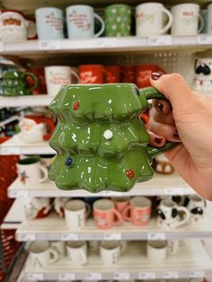 a hand holding a green ceramic christmas tree mug in front of shelves with coffee cups