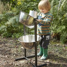 a little boy standing next to a metal bowl on a stand in the grass and plants