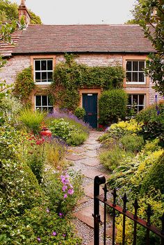 an old stone house surrounded by greenery and flowers with a gate leading to the front door