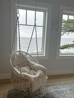 a white hanging chair in front of two windows overlooking the water and trees, with a rug on the floor