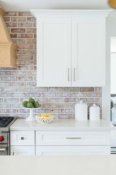 a kitchen with white cabinets and a brick wall behind the stove top is filled with fruit