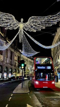 a red double decker bus driving down a street next to tall buildings covered in christmas lights