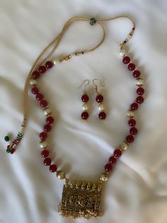 The Nashili necklace set is made with maroon and and pearl beads, with a plaque pendant depicting two handmaids fanning Ganesha in traditional Indian metalwork. The subtle details of the metalwork elevate the piece, from the borders around the plaque to the setting of the pearl beads to the structure of the necklace itself. The adjustable backing allows for some adjustments to the length of the necklace, although it is meant to hang long past the chest. Traditional Handmade Pearl Necklace For Celebration, Traditional Red Pearl Necklace For Festive Occasions, Traditional Ruby Beaded Necklace As Gift, Traditional Brass Jewelry With Polished Beads, Traditional Kundan Necklace For Gift, Festive Traditional Pearl Necklace With Polished Beads, Traditional Polished Beads Pearl Necklace For Festivals, Traditional Polished Beads Jewelry Sets As Gift, Traditional Kundan Necklace With Pearl Pendant As Gift