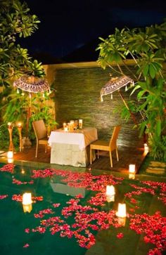 an outdoor dining area is lit up with candles and flowers on the ground next to a pool