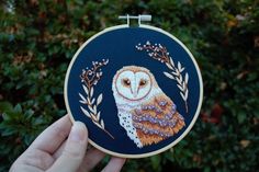 a hand holding an embroidered owl in front of some bushes