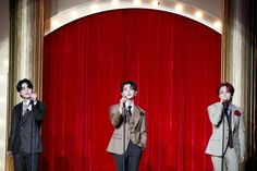 three men standing on stage with microphones in front of red curtained walls and one man wearing a suit