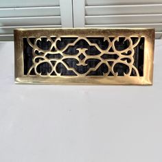 a gold metal grate on a white surface with window shutters in the background