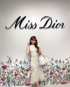 a woman standing in front of a floral wall holding a white purse and posing for the camera