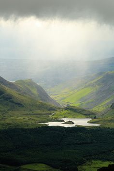 a lake in the middle of a green valley under a dark sky with storm clouds