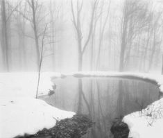 a small pond surrounded by snow covered trees in the middle of a forest with fog