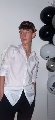 a young man wearing a white shirt and black pants with flowers in his hair standing next to balloons