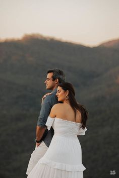 a man and woman standing next to each other on top of a hill with mountains in the background