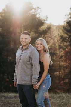 a man and woman holding hands while standing in front of some trees with the sun shining on them