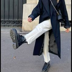 a woman in black coat and white pants walking down the street with her feet up