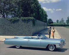 an old car parked in front of a brick wall with two people standing next to it