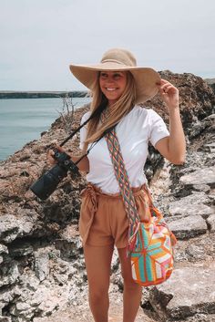 a woman wearing a hat and holding a camera in her hand while standing on rocks near the water