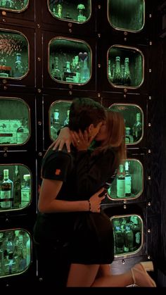 a man and woman kissing in front of many bottles on the wall with green lights behind them