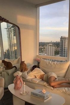 a woman laying on a couch in front of a window next to a dog and cat