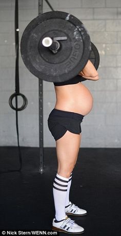 a pregnant woman lifting a barbell with no shirt on, while wearing shorts and socks