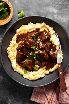 a black plate topped with mashed potatoes covered in beef and gravy next to a bowl of parsley