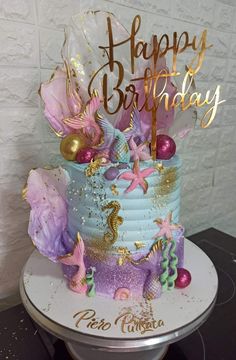 a birthday cake decorated with mermaid decorations and gold writing on it's top is sitting on a table