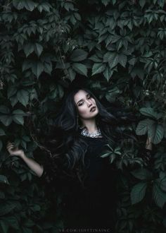 a woman with long black hair standing in front of green leaves