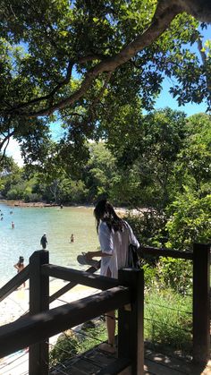 a woman standing on a wooden bridge looking out at the water and people swimming in the lake