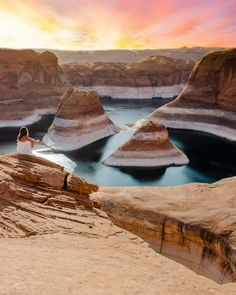 a woman sitting on top of a cliff next to a body of water at sunset