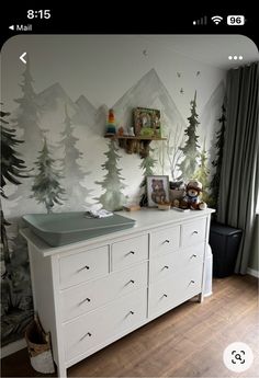 a white dresser sitting next to a window with trees on the wall and pictures above it