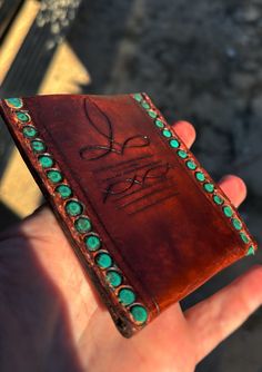 a hand holding a small brown leather wallet with green stones on it's sides