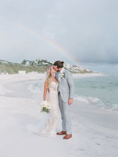 a bride and groom kissing on the beach with a rainbow in the sky behind them