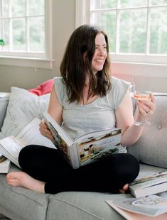 a woman sitting on a couch reading a book and holding a glass of wine in her hand