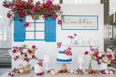 a table topped with cakes and flowers next to blue shuttered window sill filled with pink roses