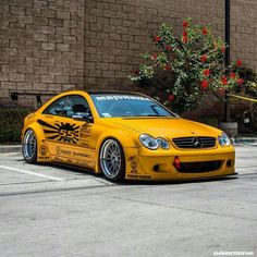 a yellow car parked in a parking lot next to a brick wall and red flowers