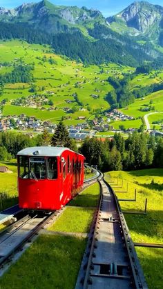 a red train traveling through a lush green hillside covered in trees and grass with mountains in the background