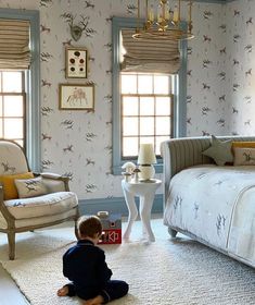 a small child sitting on the floor in front of a bedroom with two windows and a bed