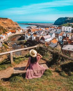 a woman in a hat sitting on a bench looking out at the ocean and buildings