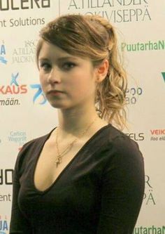 a young woman standing in front of a white wall wearing a black shirt and gold necklace