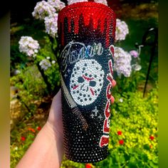 a hand holding a red and black bottle with a face painted on it in front of some flowers