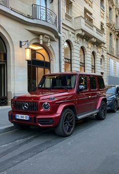 a red mercedes g - class is parked on the street in front of a building