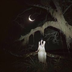 a woman in white dress standing under a tree at night with the moon behind her