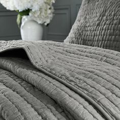 a bed with a gray comforter and white flowers in a vase on the side