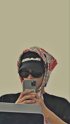 a woman wearing sunglasses and a headscarf is looking at her cell phone while sitting in front of a laptop