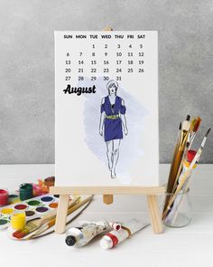 a calendar with an image of a woman's dress on it next to paintbrushes and watercolors