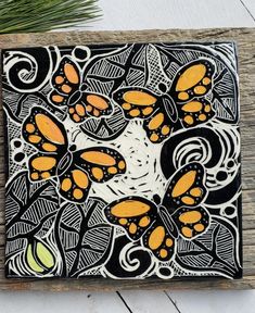 an art piece with orange and black designs on it sitting on a wooden surface next to a pine tree